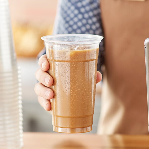 A person holding a Choice Heavy Weight clear plastic cup of brown liquid.