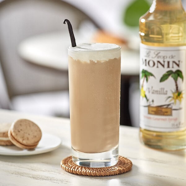A glass of milkshake made with Monin Premium Vanilla Flavoring Syrup on a coaster with a cookie and a bottle of syrup.