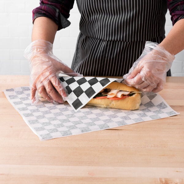 A person wearing gloves and using Choice black check deli wrap paper to wrap a sandwich with meat and vegetables.