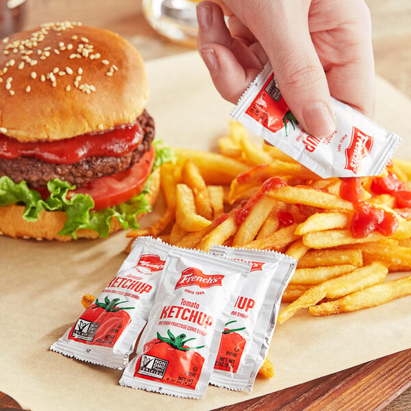 A person holding a French's tomato ketchup packet and putting it on a hamburger.