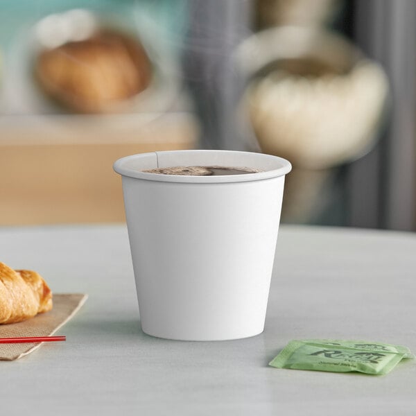 A white Choice paper hot cup filled with brown liquid on a table with a croissant