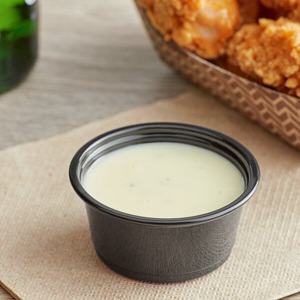 A bowl of white sauce next to fried chicken wings served in a black plastic souffle cup.