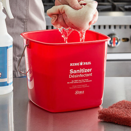 A person's hand washing a red sponge in a red Noble Products sanitizing pail on a counter.