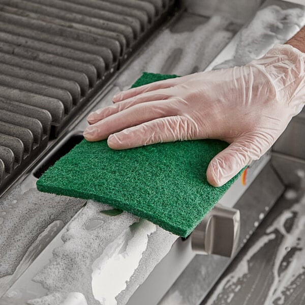 A person wearing gloves using a Lavex green scouring pad to clean a grill.